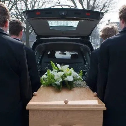 Porteurs de cercueil en costume chargeant un cercueil en bois orné de fleurs dans un corbillard.
